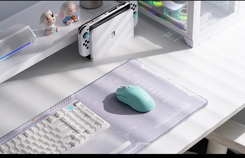 Teal mouse on a desk with a keyboard, gaming console, and laptop in the background.