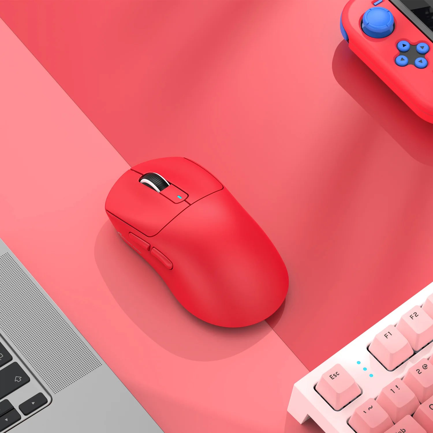 Red computer mouse on a pink surface with a laptop and keyboard in the background